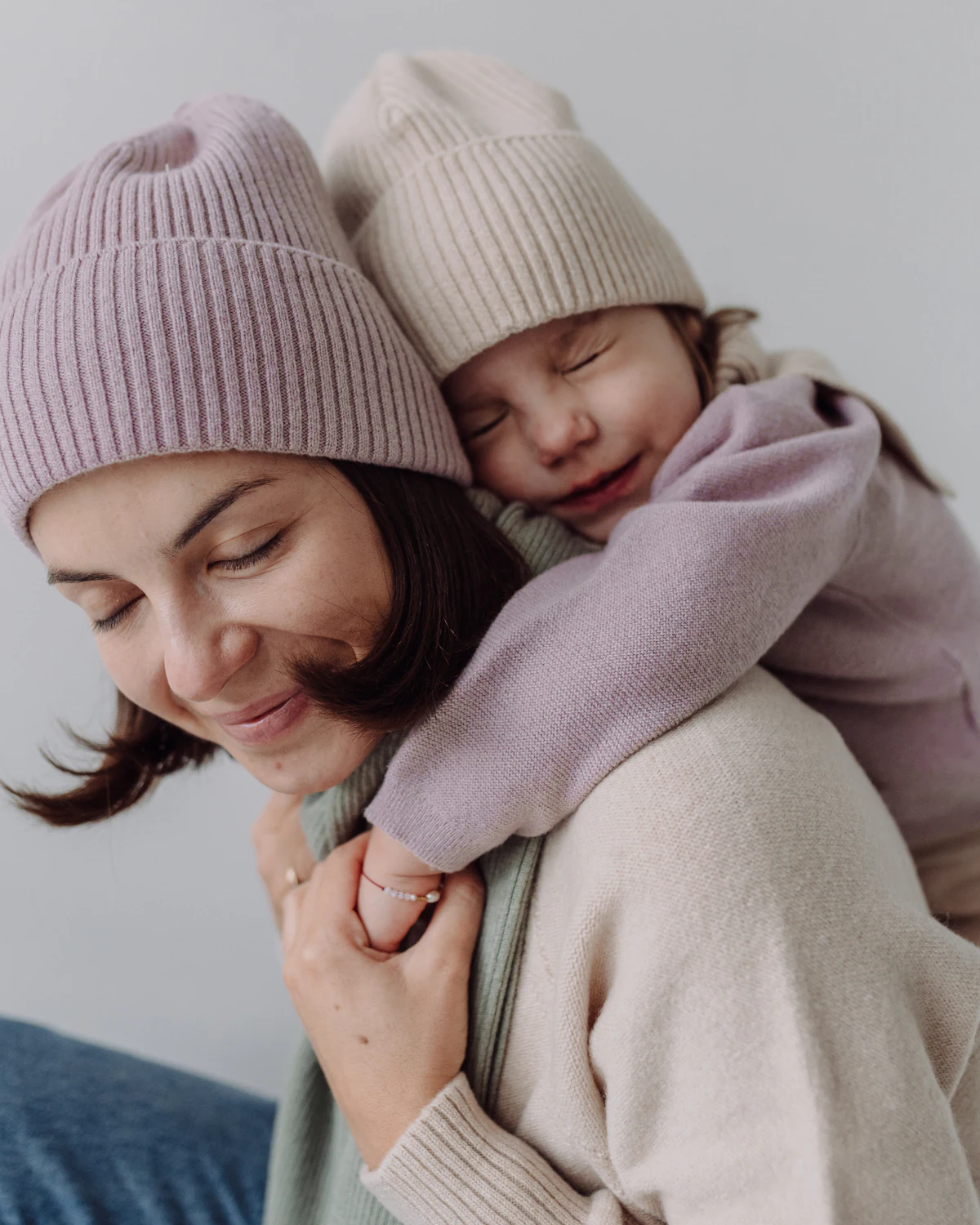 Mama w różowej czapce z wełny merino niesie na plecach córkę w beżowej czapce
