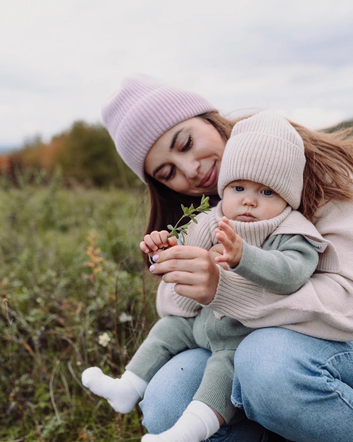 Mama z niemowlęciem w czapkach i kominie z wełny merino na łące