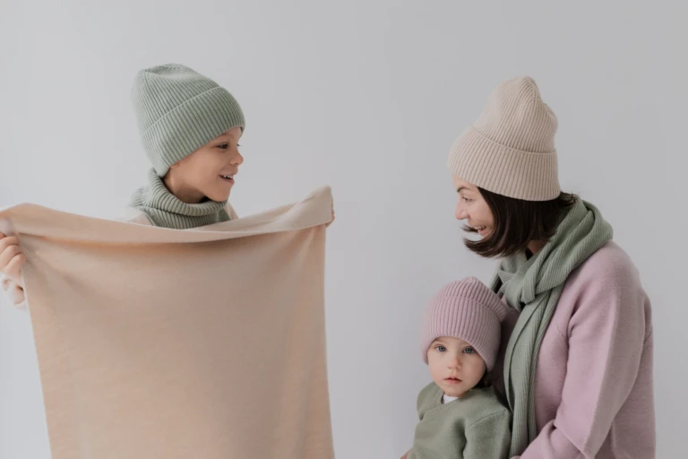 Chłopiec trzyma beżowy kocyk z wełny merino, obok mama z córką w czapkach