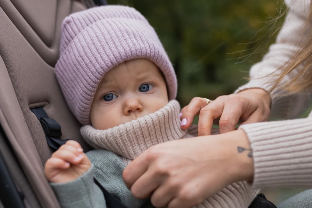 Niemowlę w różowej czapce i kominie z wełny merino w wózku, mama poprawia mu komin