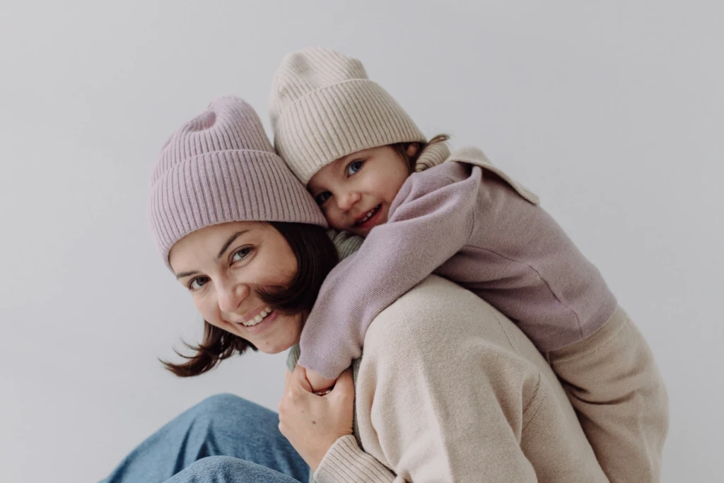 Mama w różowej czapce z córką na plecach, obie w czapkach i swetrach z wełny merino