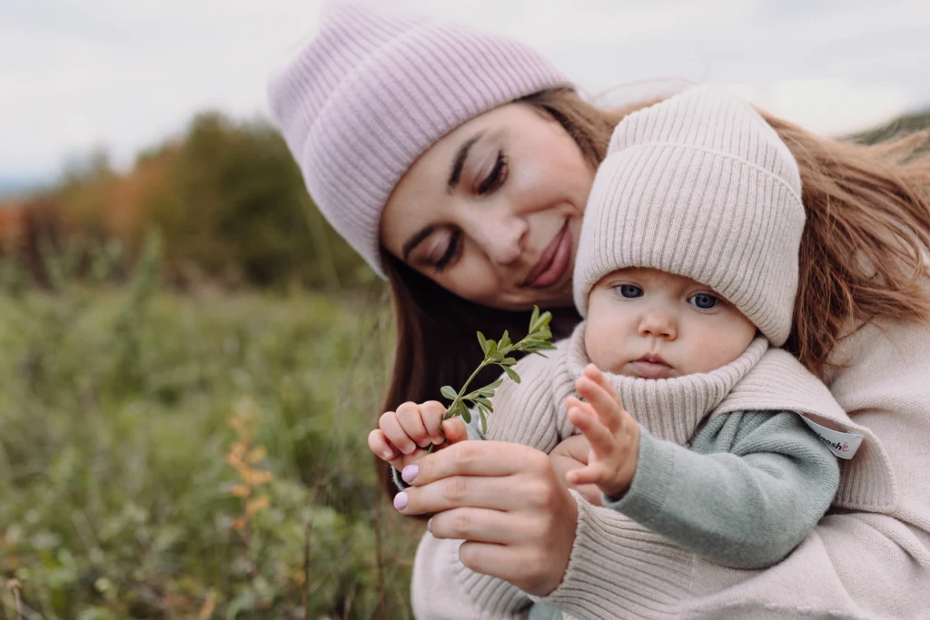 Mama i niemowlę w czapkach z wełny merino oglądają gałązkę na jesiennej łące
