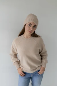 Kobieta w beżowej czapce merino dla dorosłych i swetrze, z dłońmi przy kieszeniach jeansów, na jasnym tle