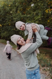 Mama w beżowej czapce merino dla dorosłych podrzuca malucha na ścieżce, w tle biegnie starszy syn