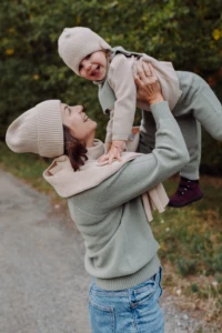 Roześmiana mama w beżowej czapce merino dla dorosłych podrzuca malucha, zbliżenie z profilu