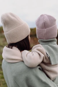 Mama w beżowej czapce merino dla dorosłych i szaliku niesie malucha w różowej czapce, widok od tyłu