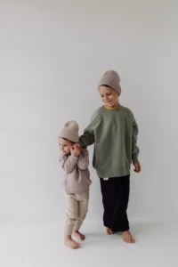 Maluch w beżowej czapce merino dziecięcej trzyma brata za rękę, obaj boso na jasnym tle