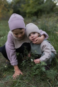 Maluch w beżowej czapce merino dziecięcej kuca w trawie, starszy brat obejmuje go ramieniem