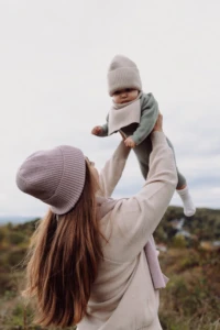 Kobieta podrzuca niemowlę w beżowej czapce merino niemowlęcej i kominie na tle nieba