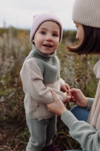 Uśmiechnięty maluch w beżowym kardiganie merino dziecięcym trzyma mamę za ręce na łące