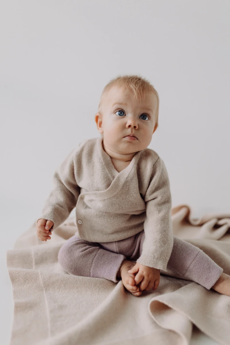 Niemowlę w beżowym kardiganie merino dziecięcym siedzi na kocyku i patrzy w górę