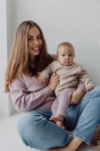 Uśmiechnięta mama trzyma na kolanach niemowlę w beżowym kardiganie merino dziecięcym