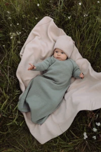 Niemowlę z wyciągniętą rączką leży na beżowym kocyku merino w trawie, widok z góry