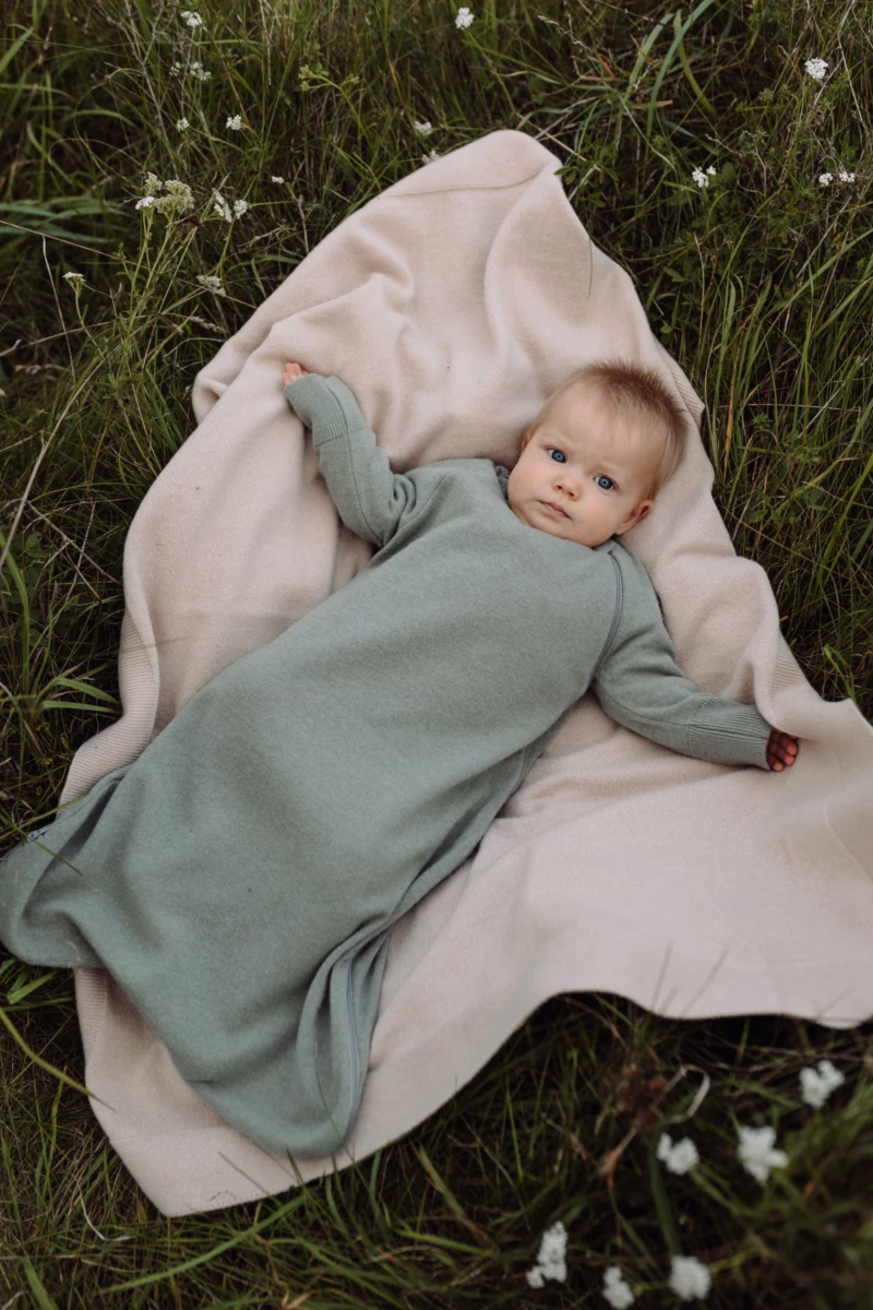 Niemowlę w zielonym śpiworku leży na beżowym kocyku merino rozłożonym na trawie, widok z góry