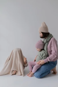 Chłopiec schowany pod beżowym kocykiem merino, obok mama z córką na kolanach