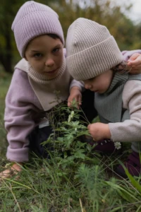 Chłopiec w beżowym kominie merino dziecięcym i maluch kucają przy roślinie w trawie