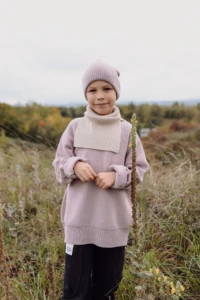 Chłopiec w beżowym kominie merino dziecięcym stoi na jesiennej łące obok wysokiej rośliny