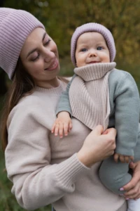 Mama w różowej czapce z uśmiechem patrzy na niemowlę w beżowym kominie merino niemowlęcym
