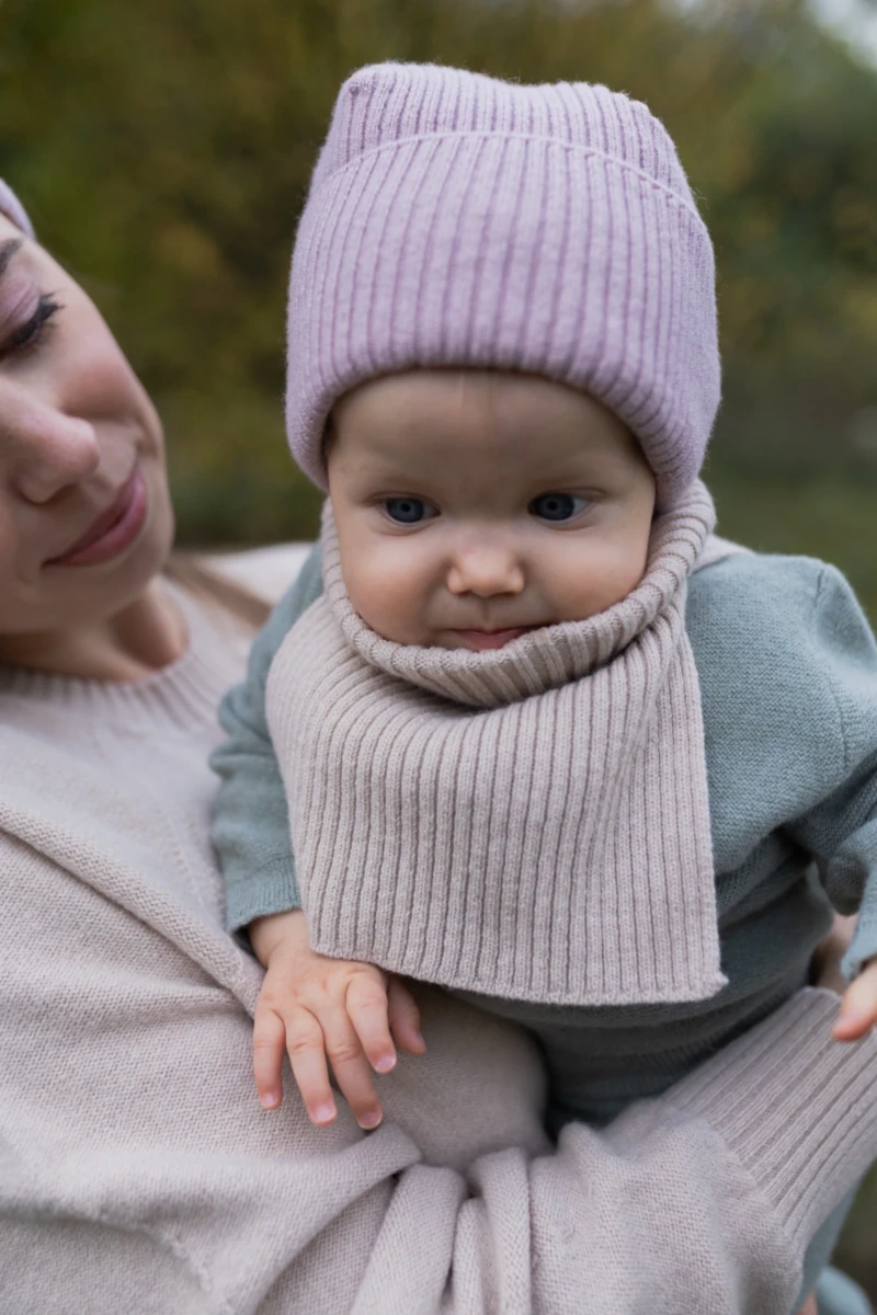 Niemowlę w beżowym kominie merino niemowlęcym i różowej czapce na rękach mamy, zbliżenie