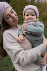 Mama w beżowym swetrze merino damskim i różowej czapce trzyma uśmiechnięte niemowlę