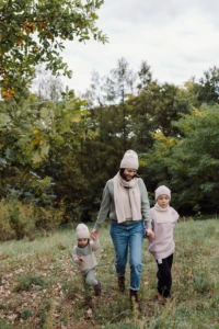Mama w beżowym szalu merino idzie przez łąkę z dwójką dzieci za rękę