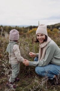 Mama w beżowym szalu merino kuca na łące i trzyma malucha za rękę