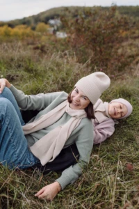 Mama w beżowym szalu merino leży na trawie z synem i uśmiecha się do obiektywu
